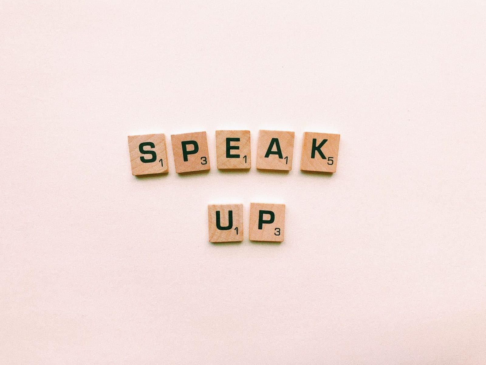 Wooden Scrabble tiles spelling 'Speak Up' on a pink background.