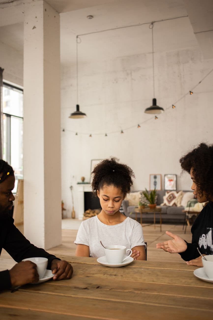 Dissatisfied African American mother gesticulating while scolding upset daughter sitting at table near crop father