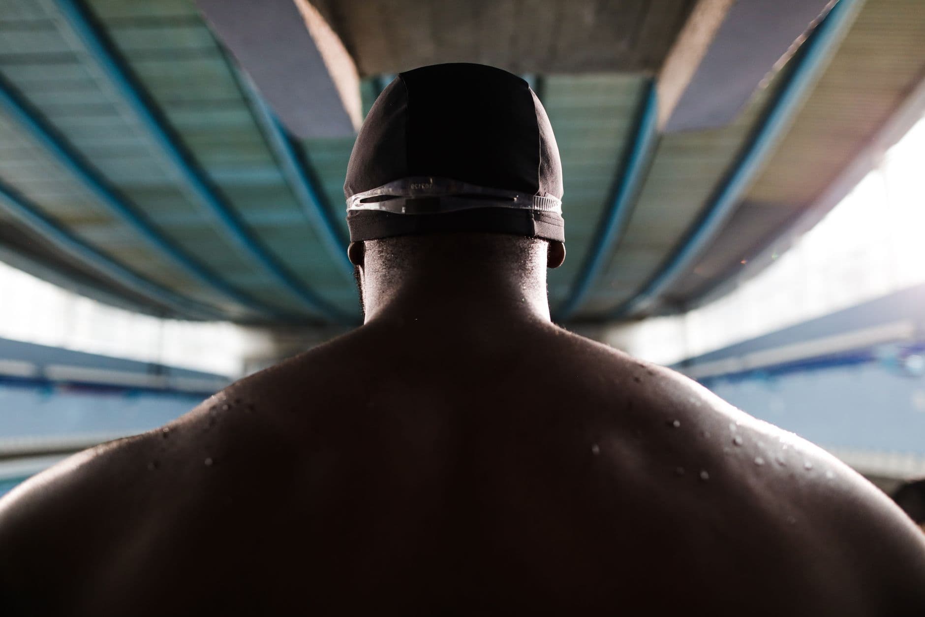 Back view of a shirtless swimmer with a cap, water droplets on skin, indoors.