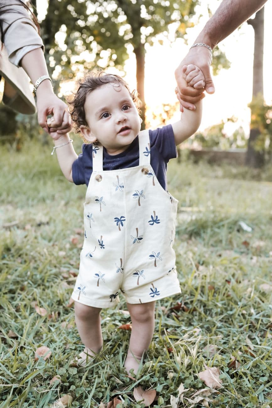 Cute baby with parents' support taking steps on a sunny day in the park.
