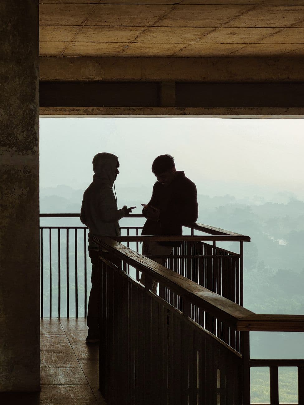 Two men in silhouette converse on a balcony with a misty background, creating a moody and introspective scene.