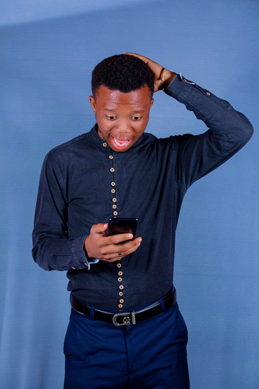 A young man in a black shirt looks surprised as he checks his smartphone against a blue background.