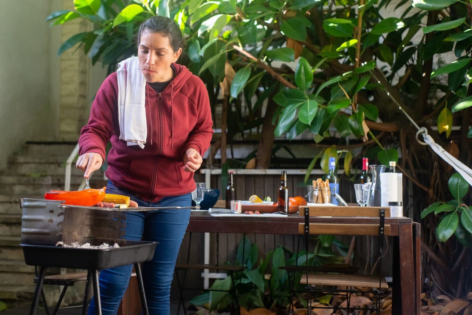 Woman grilling food outdoors, surrounded by plants and barbecue tools. Perfect for lifestyle and food themes.