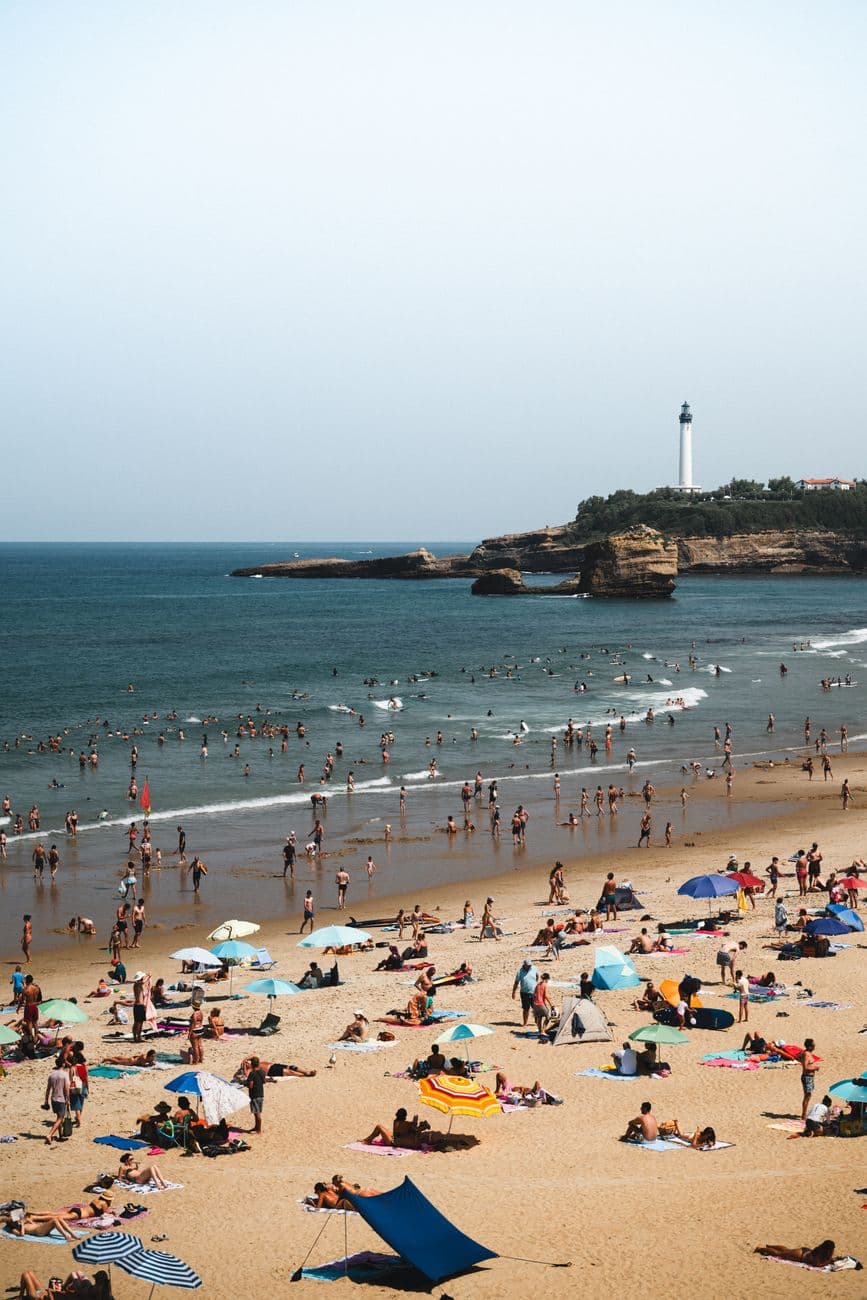 A vibrant summer scene of a crowded beach in Biarritz with a distant lighthouse.