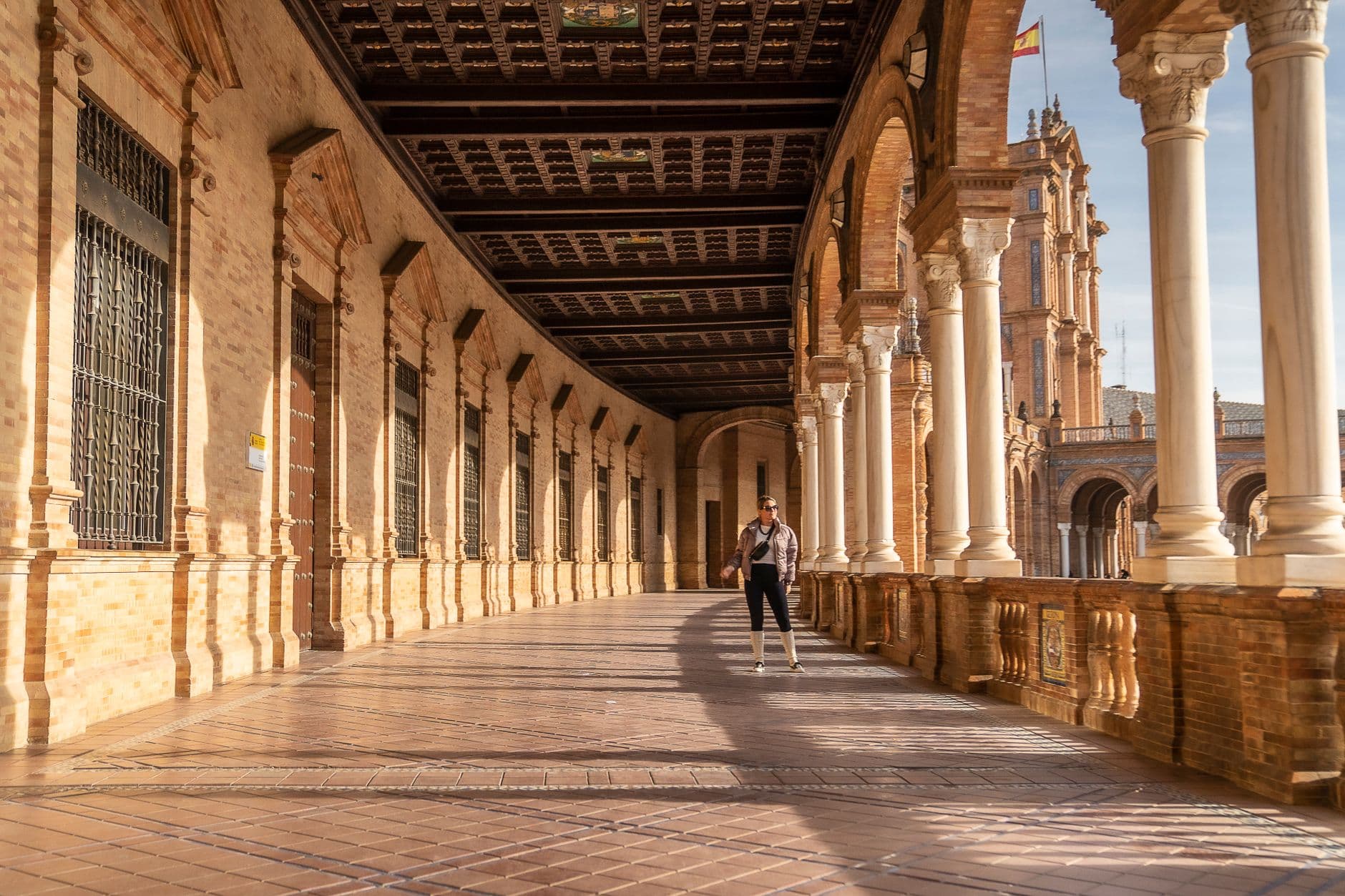 A person stands under the sunlit arches of Plaza de España in Seville, Spain.