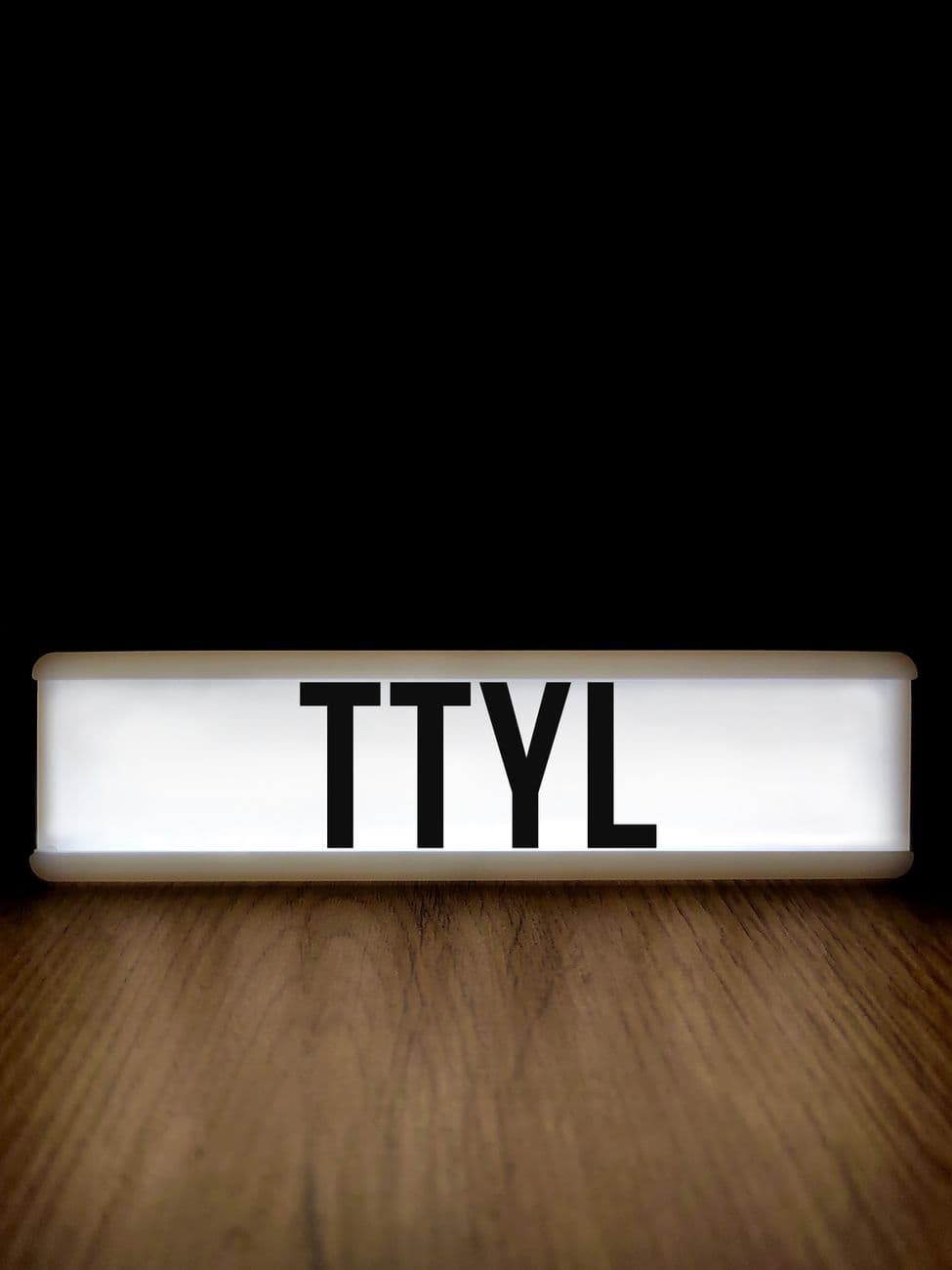 Close-up of a lightbox displaying the text TTYL, set on a wood surface against a black background.