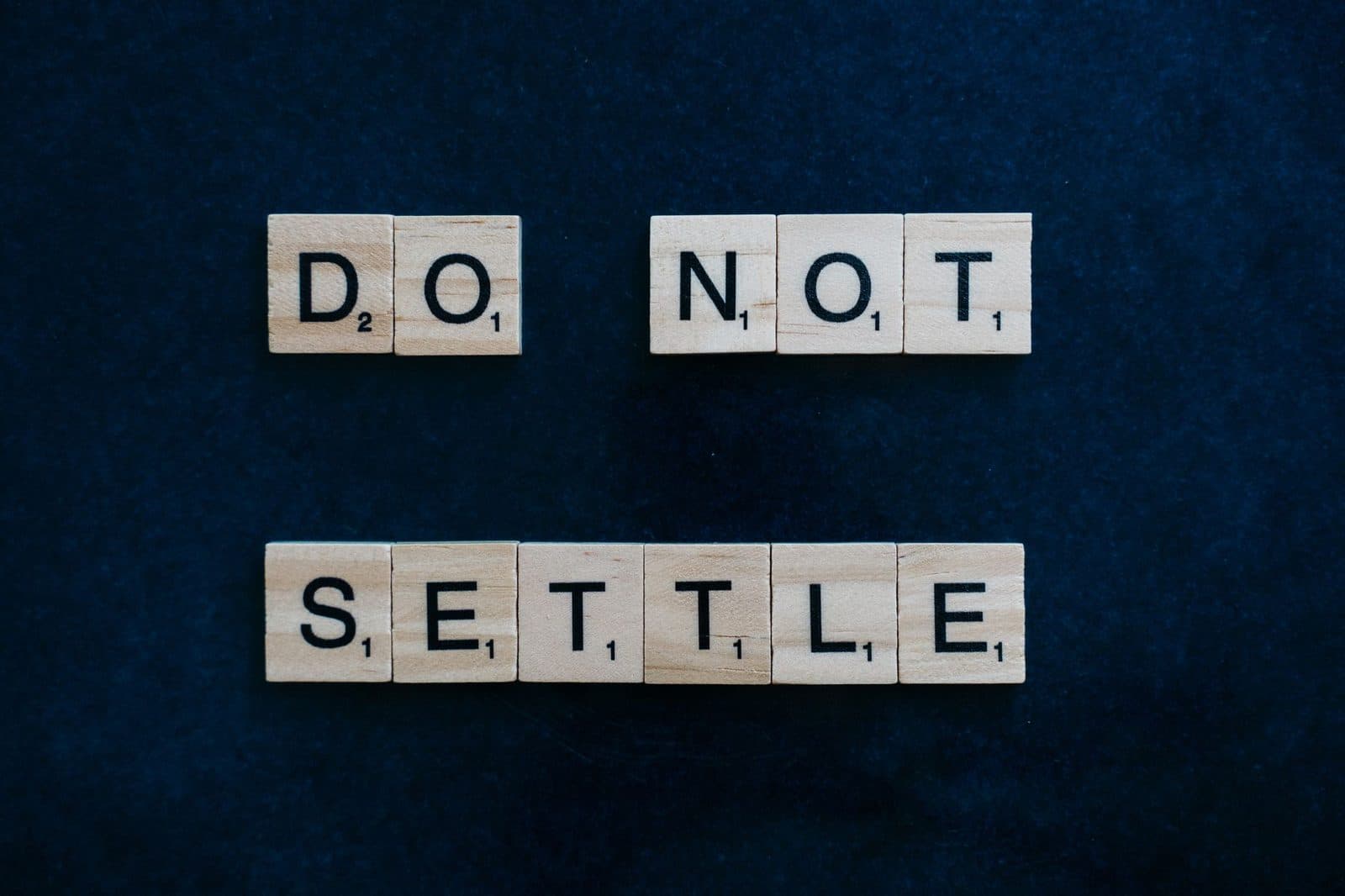 Wooden Scrabble tiles spelling 'Do Not Settle' on a dark surface, top view.