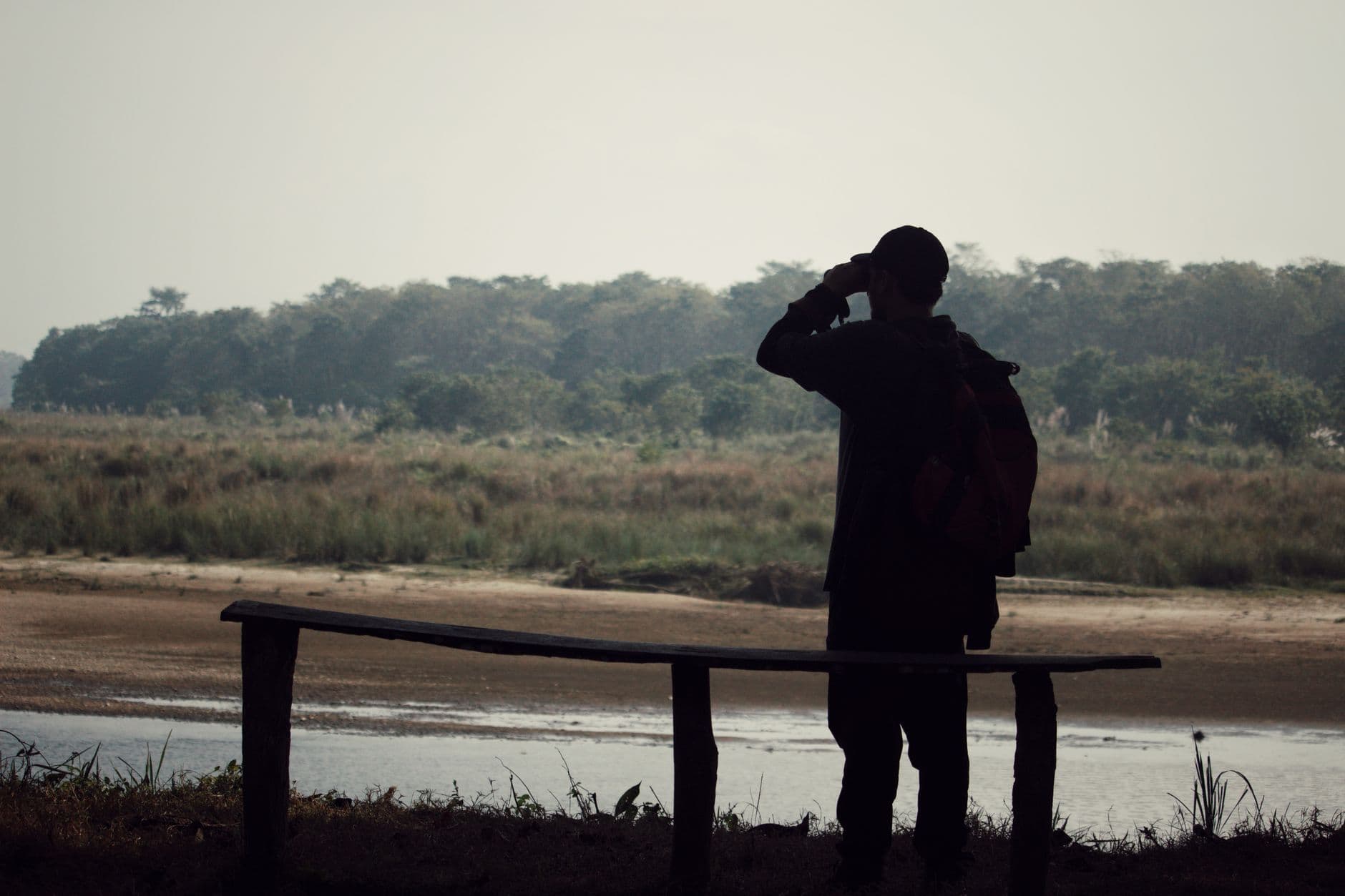 A man with a backpack observes a marsh landscape using binoculars, standing near water.