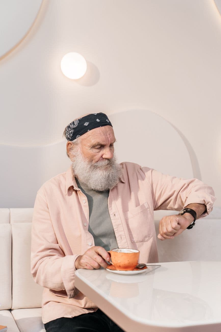 Senior man in casual wear with beard and bandana looks at wristwatch while holding mug at cafe.