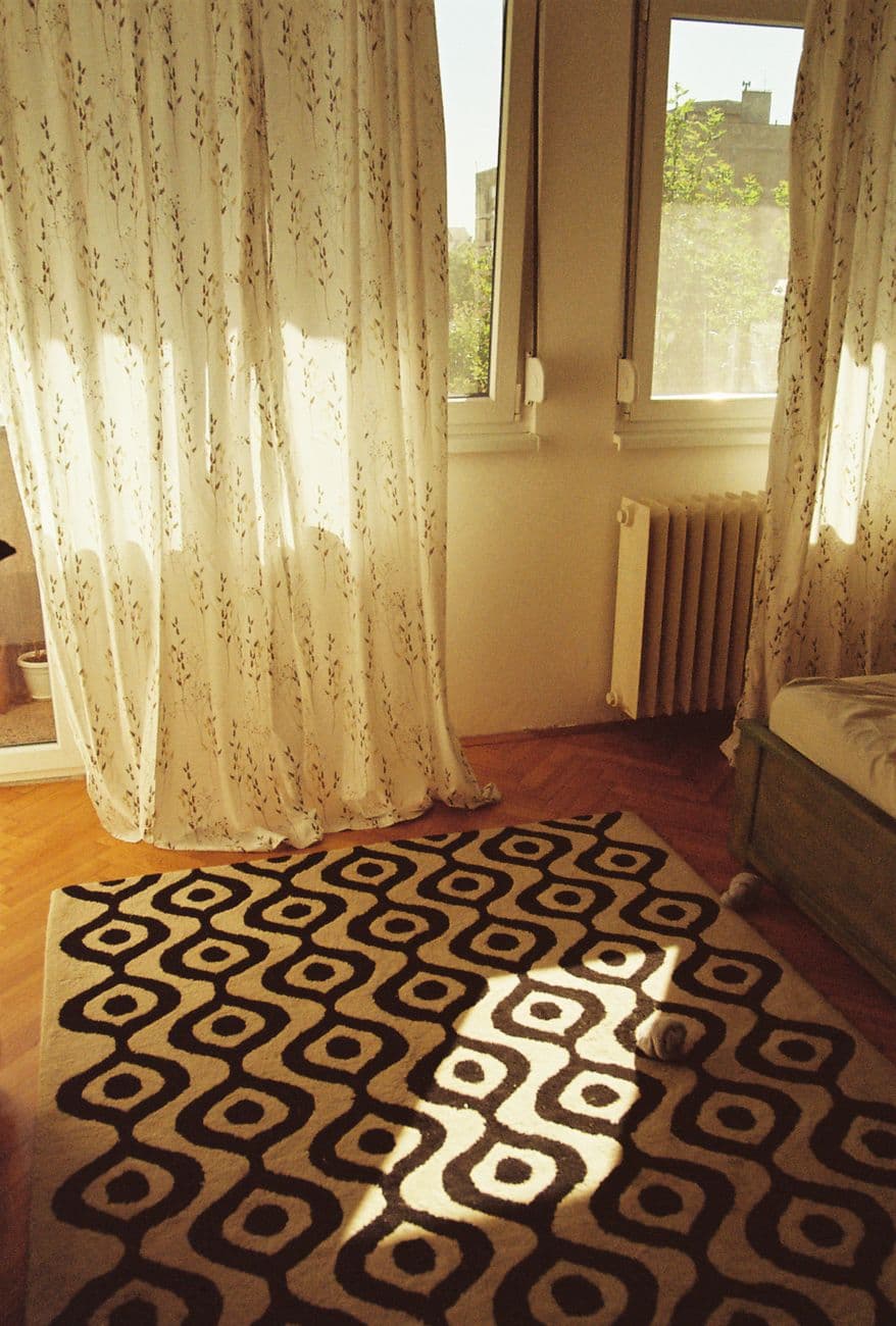 Warm and inviting bedroom with sunlight streaming through lace curtains and a stylish geometric rug.