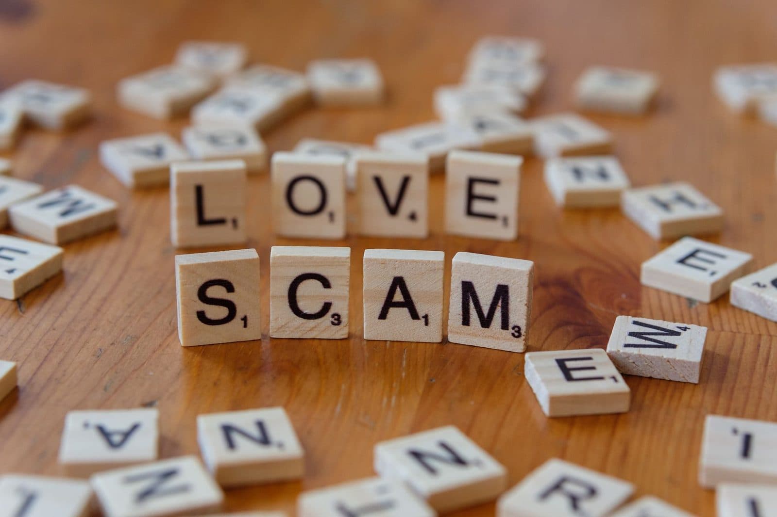 Scrabble tiles form words 'LOVE' and 'SCAM' on a wooden surface, illustrating deceit in relationships.