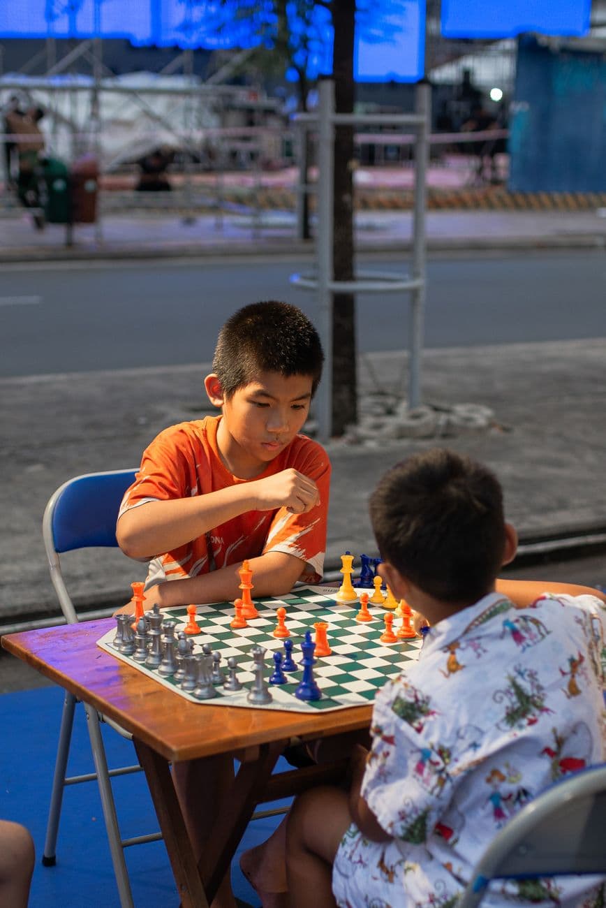 Two young boys intensely focus on a chess match outdoors, surrounded by urban evening ambiance.