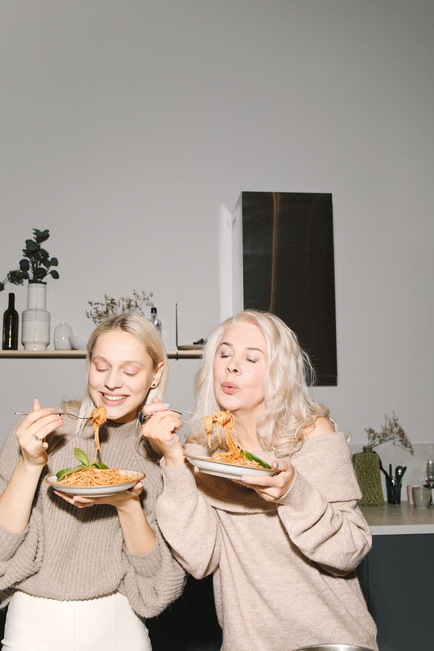 A joyful mother and daughter enjoying spaghetti together in a cozy kitchen.