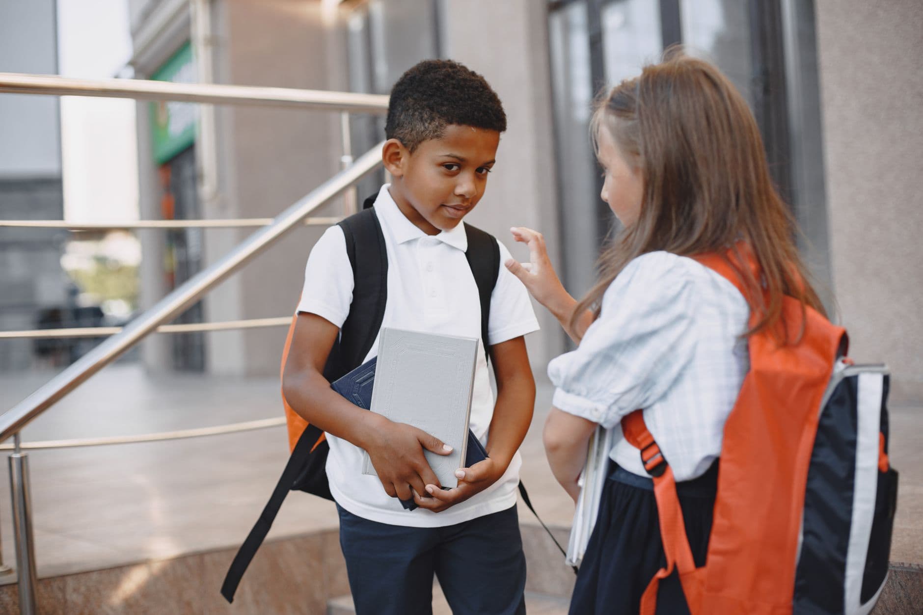 Two children with backpacks conversing outside a school building.