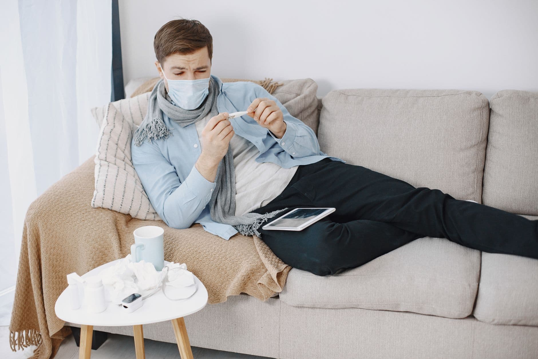 Man wearing a mask checks a thermometer while resting on a couch, surrounded by tissues.