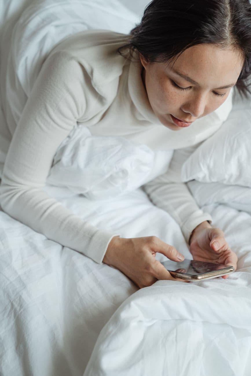 A woman in a white sweater uses her smartphone while relaxing in bed, enjoying leisure time indoors.