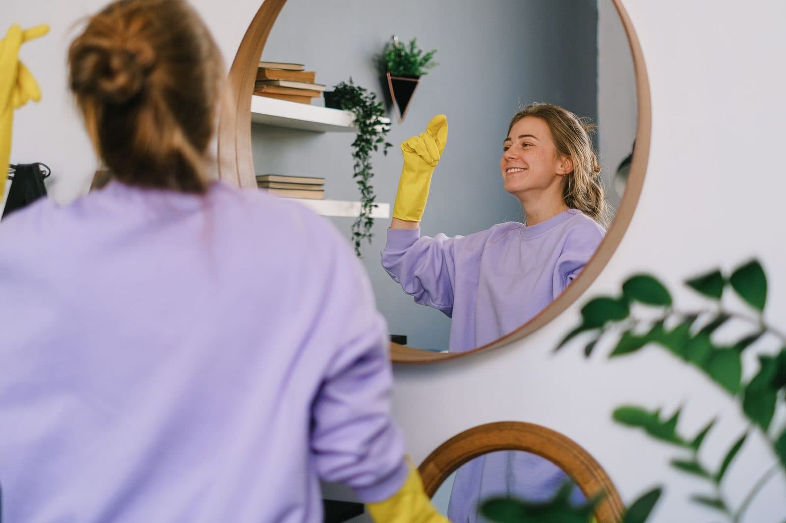 Smiling woman cleaning mirror with yellow gloves indoors, reflecting her image.