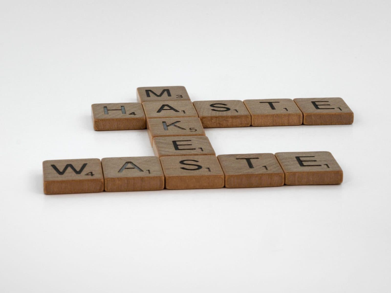Wooden Scrabble tiles spelling 'Haste Makes Waste' arranged on a white surface, symbolizing patience.
