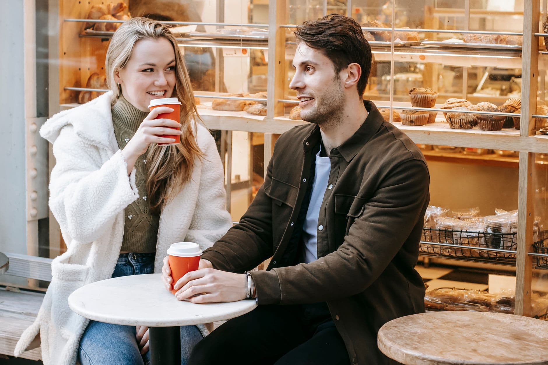 Delighted young female with long hair and unshaven male in jacket having date with takeaway coffee in outdoors cafeteria