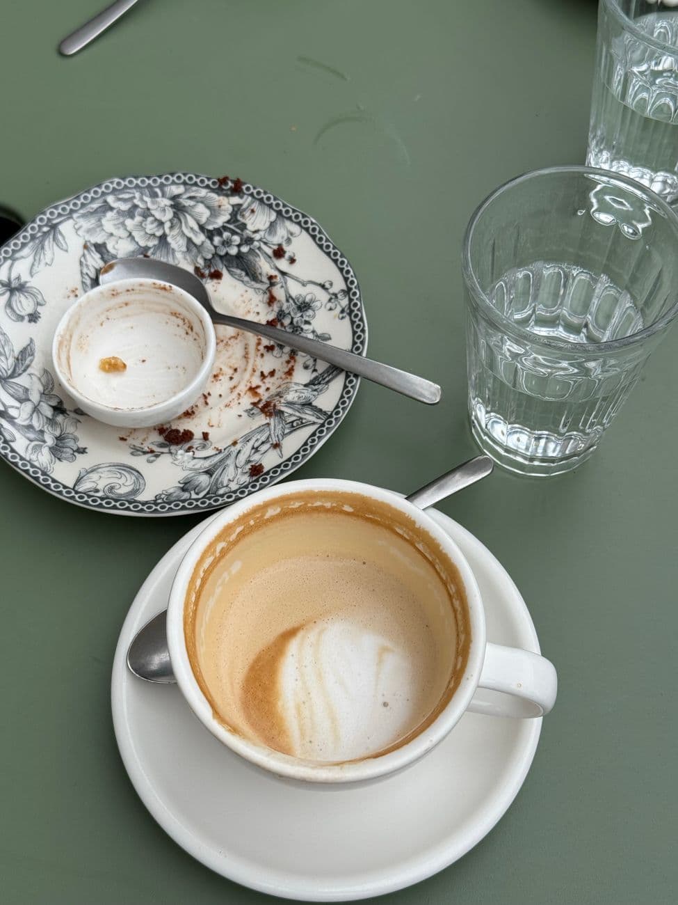 A close-up of a half-drunk cappuccino and empty dessert plate with glass cups on a cafe table.