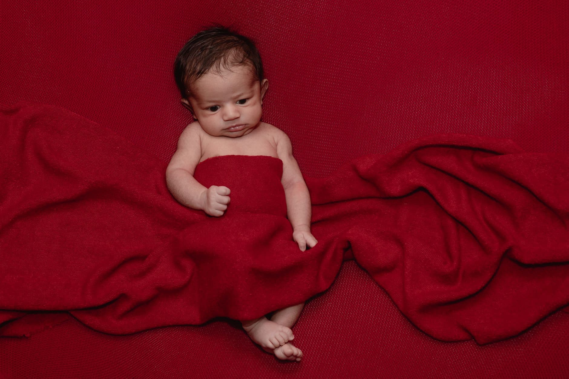 Adorable newborn baby wrapped in a red blanket, lying on red fabric.
