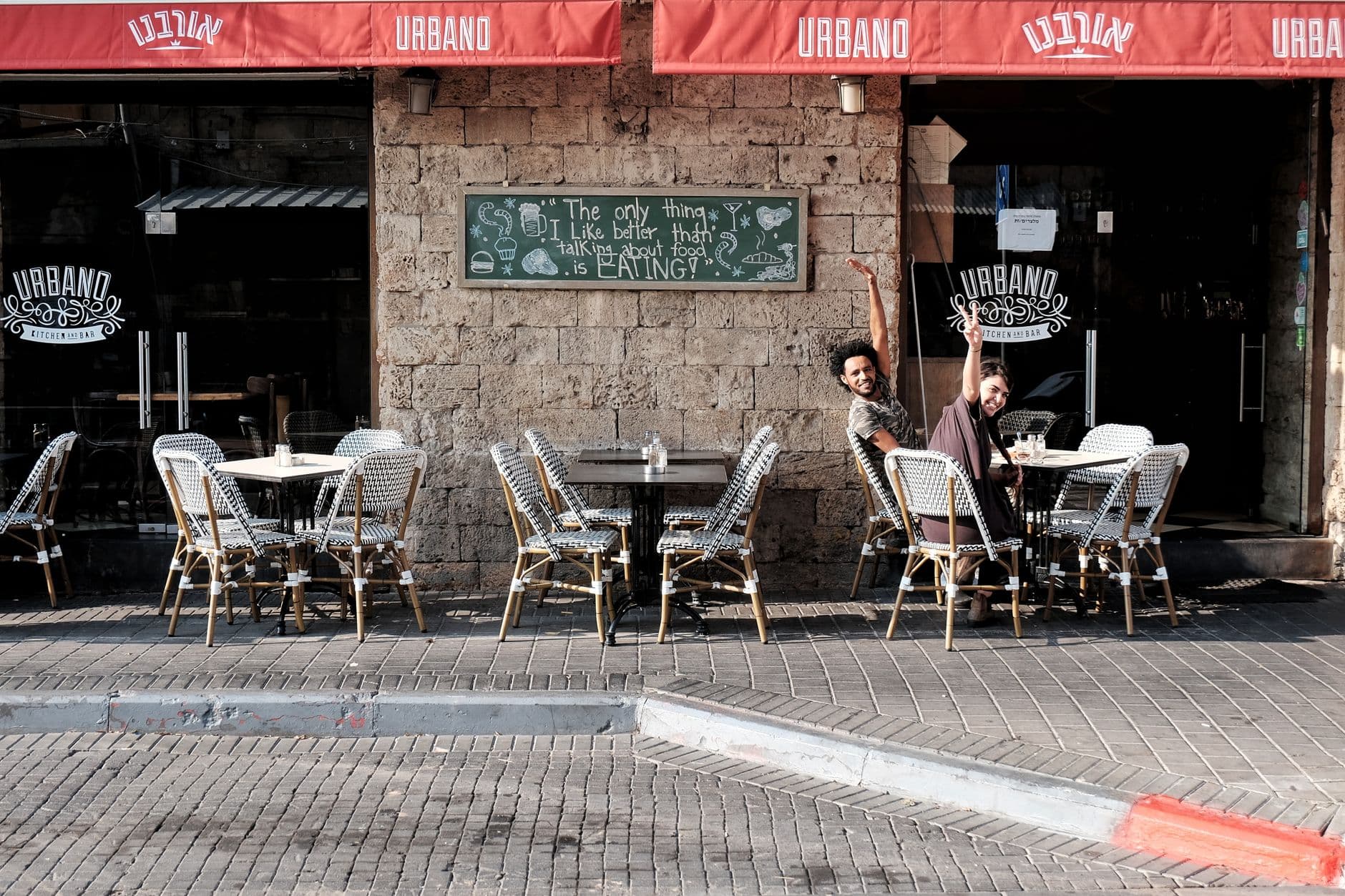Two people joyfully pose at a sunlit sidewalk cafe named Urbano, adding vibrancy to the urban setting.