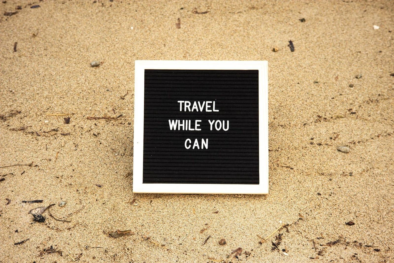 Quote 'Travel While You Can' on board in sand, inspiring travel and adventure.