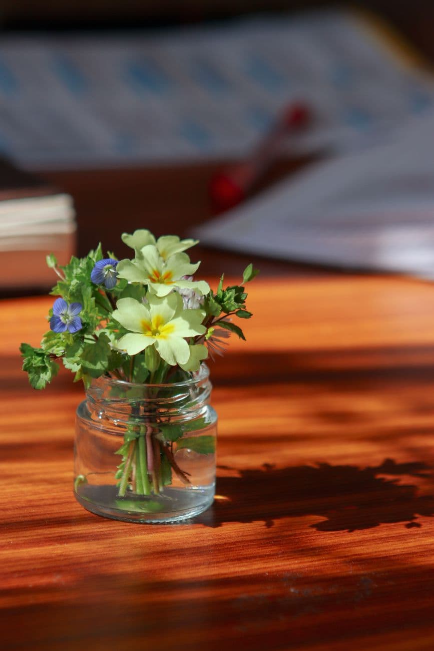 A small bouquet of wildflowers in a glass jar casting a shadow on a wooden table.