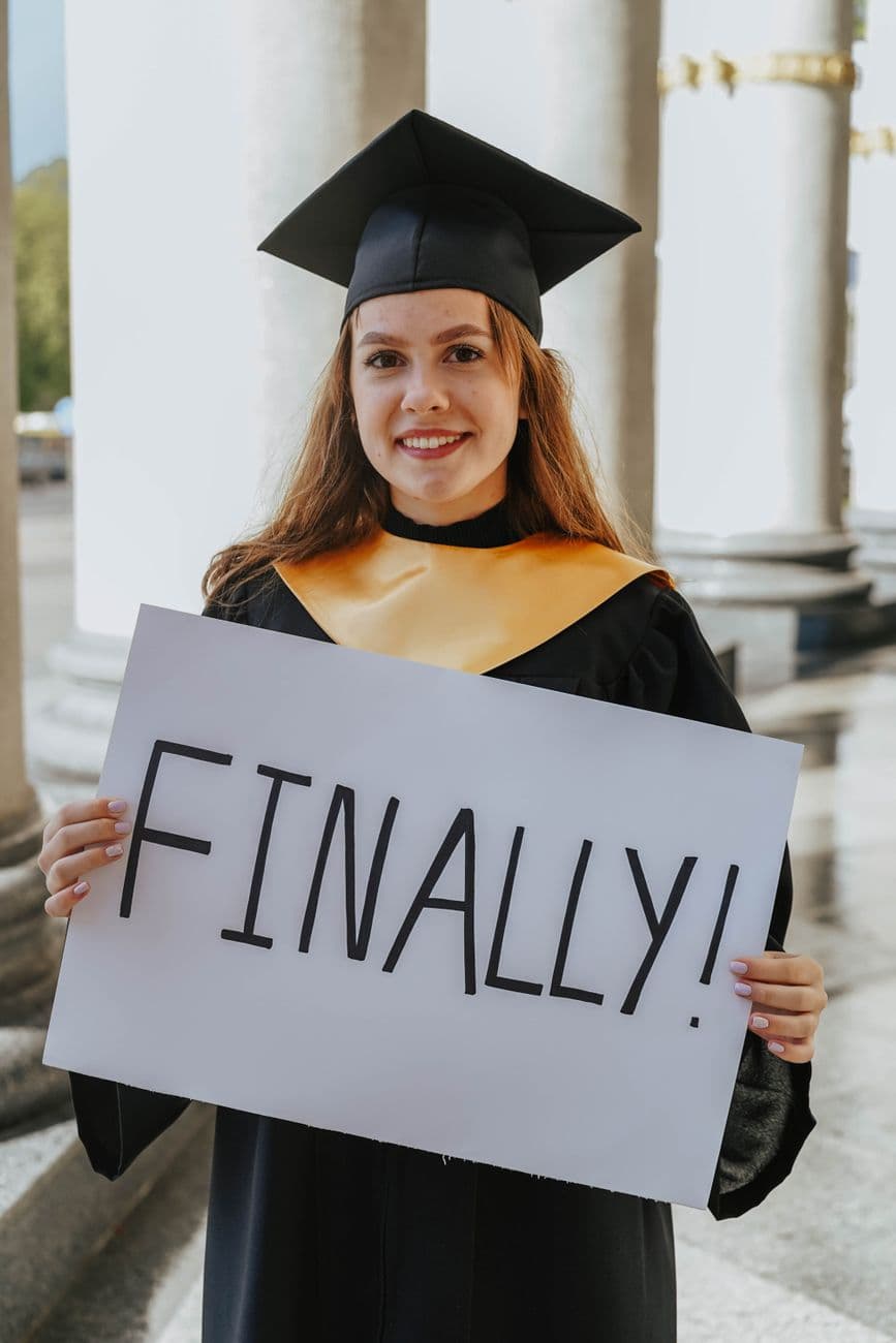 A joyful young woman in a graduation gown holding a sign that reads 'Finally!' outdoors.
