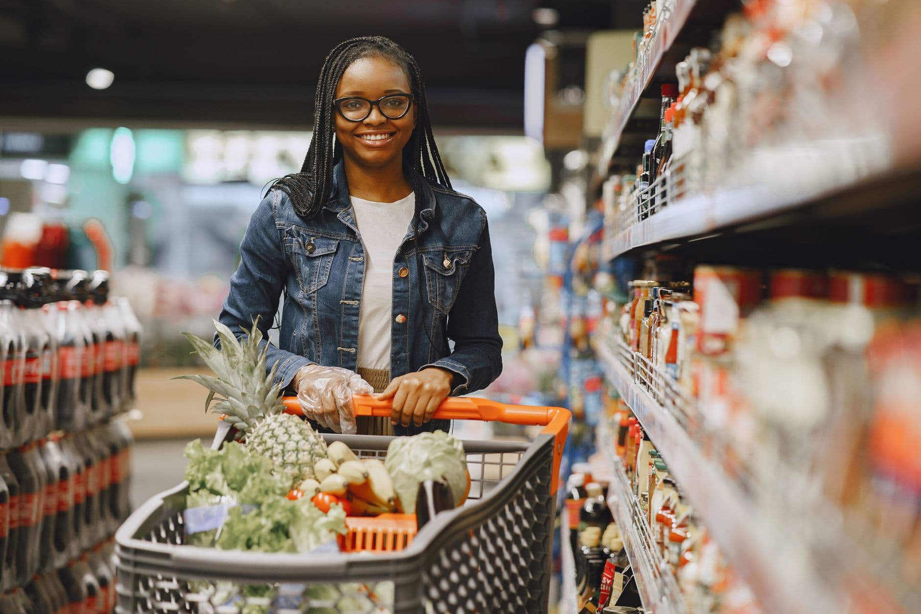 African American woman with glasses shopping in a grocery store aisle with a full cart of fresh produce.