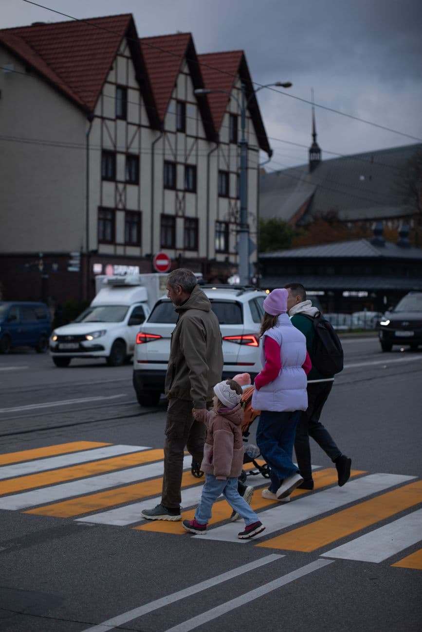 A family crosses a street at dusk in an urban area with vehicles and buildings.