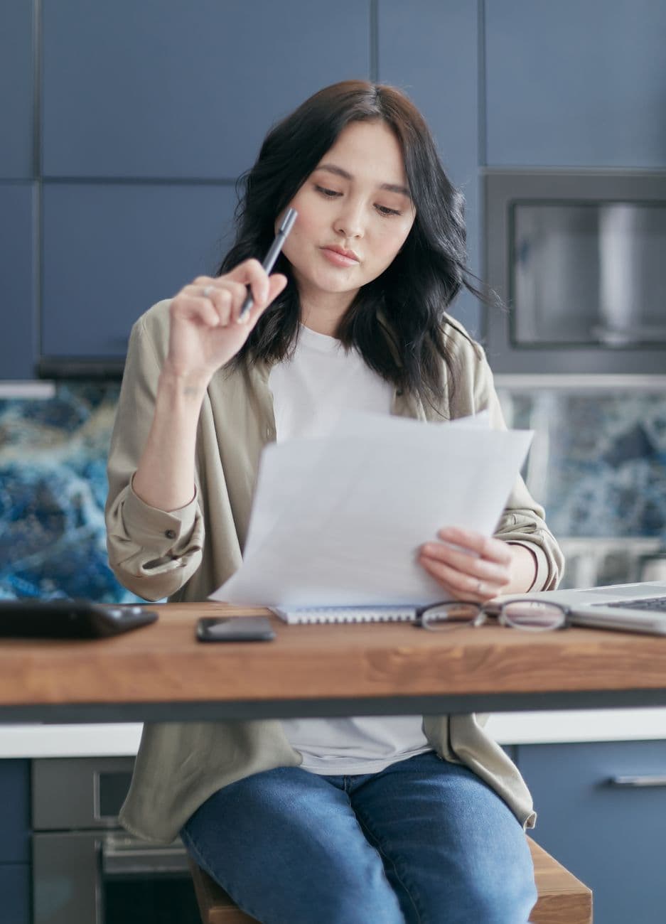 Woman sitting at a kitchen counter, pensively reviewing documents.