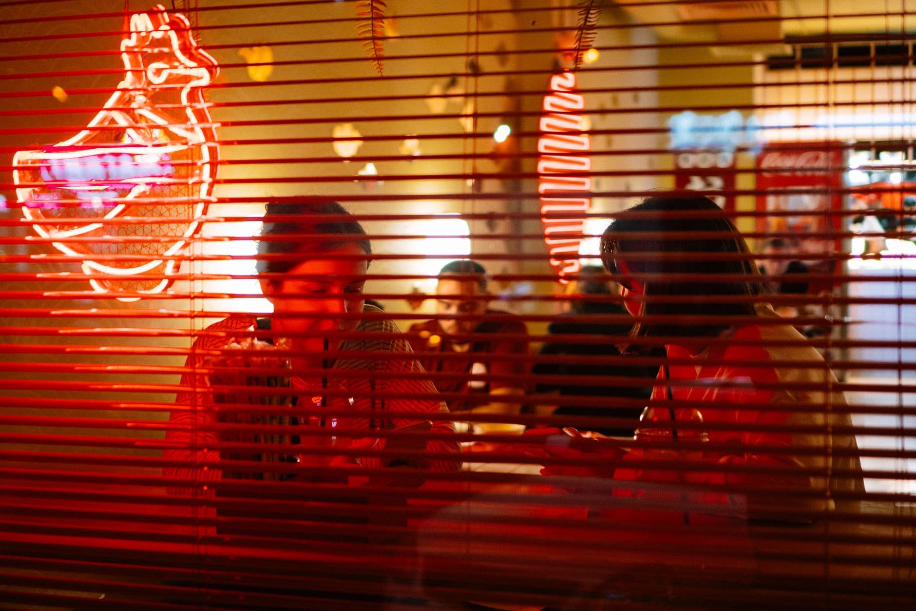 Two women enjoying dinner inside a neon-lit restaurant, viewed through red blinds.