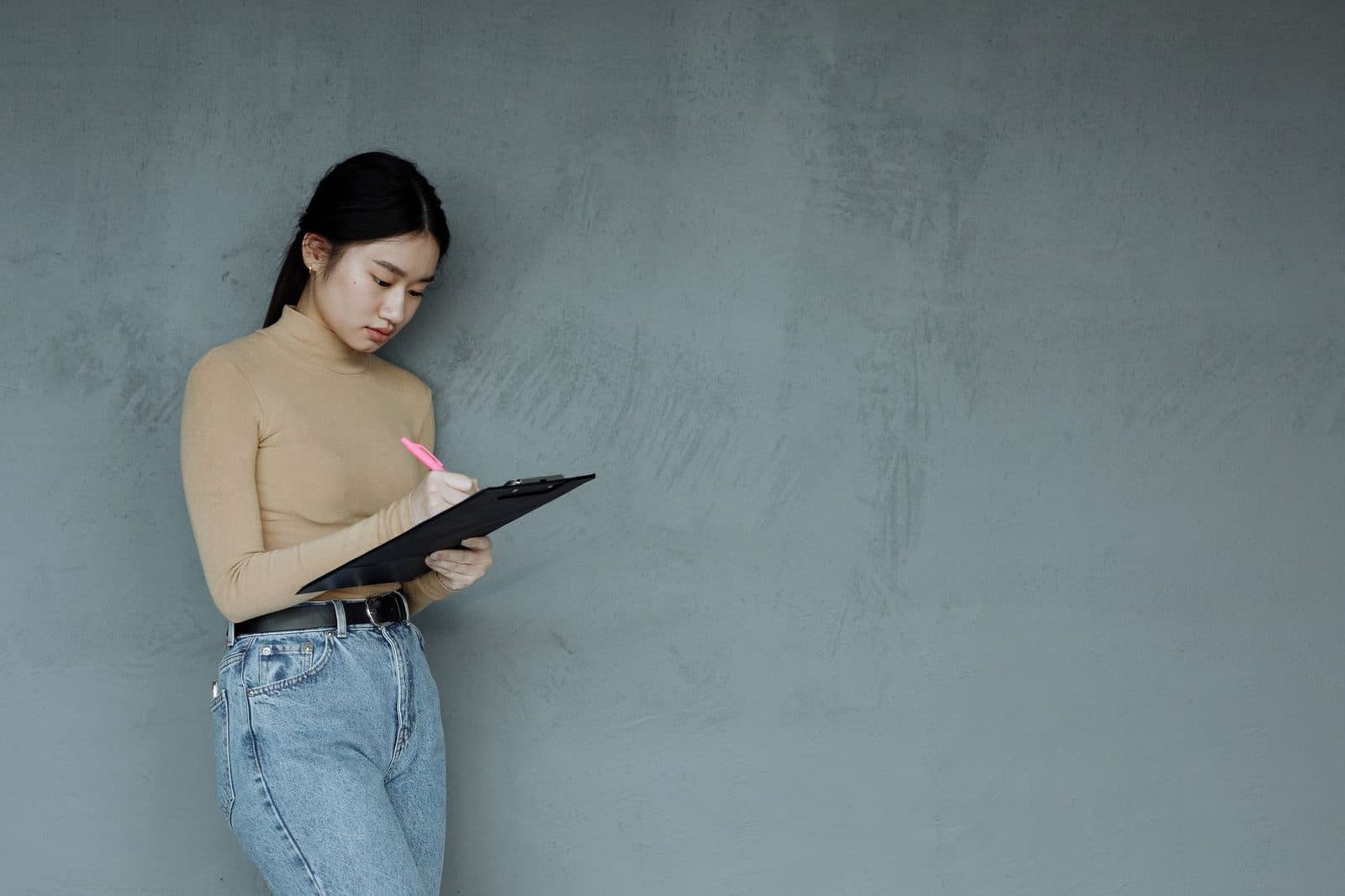 Asian woman writing notes on a clipboard standing against a minimalist gray wall.