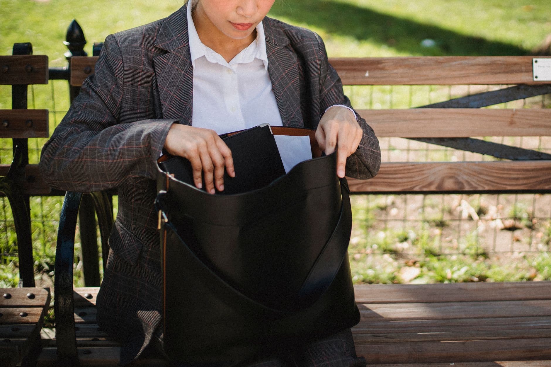 A woman in a suit searching her bag while sitting on a park bench on a sunny day.