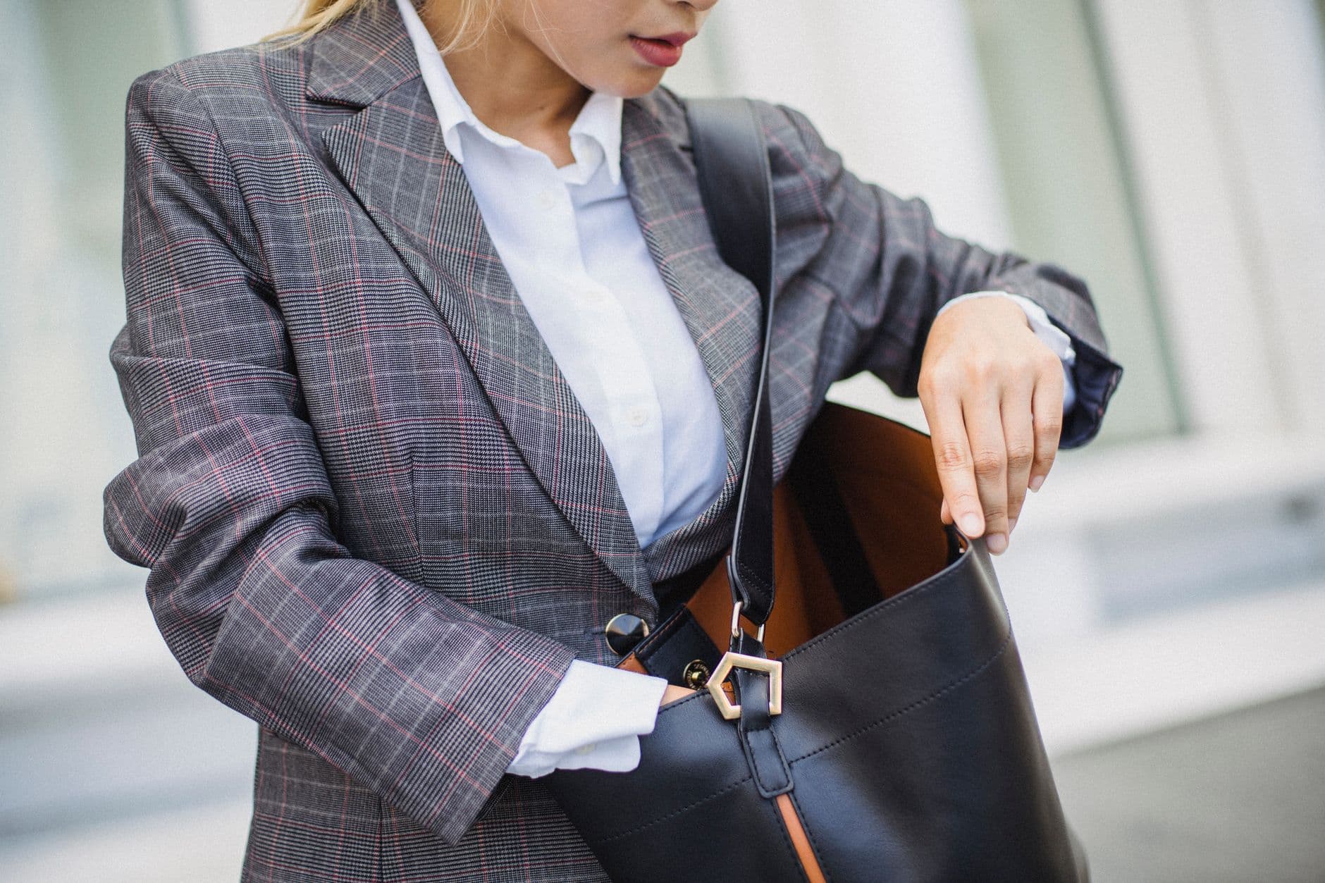 Close-up of a woman in a plaid blazer searching inside a leather bag outdoors.