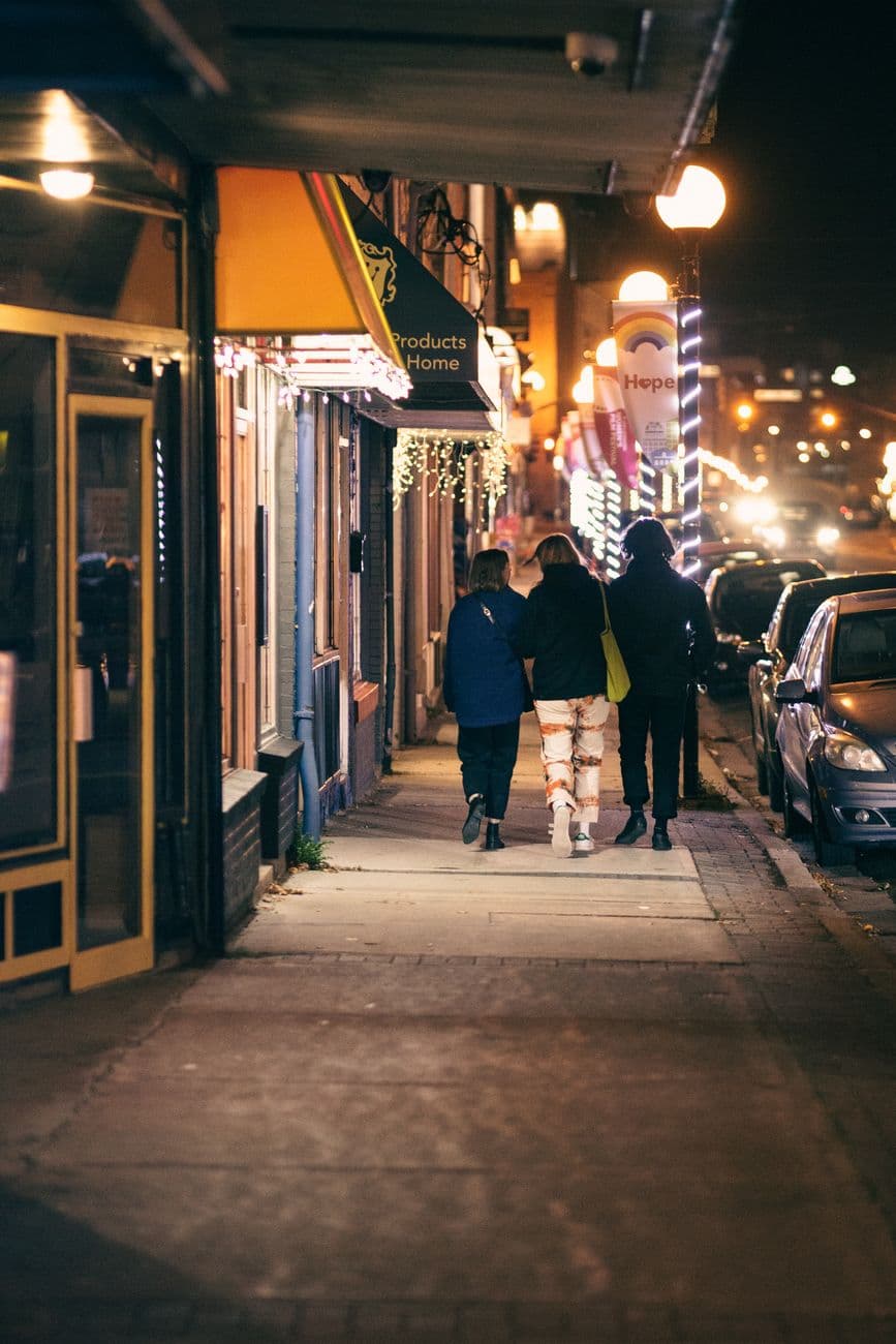 Back view of anonymous partners in outerwear strolling on walkway between buildings and roadway with shiny lights in evening city
