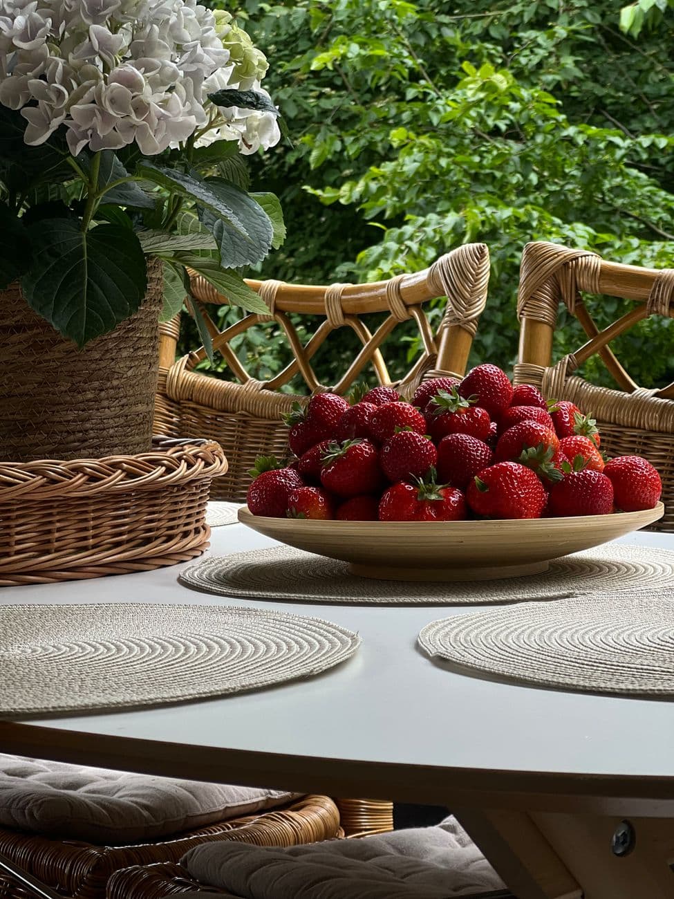 A bowl of fresh strawberries on a white table with wicker chairs and a potted plant outdoors.