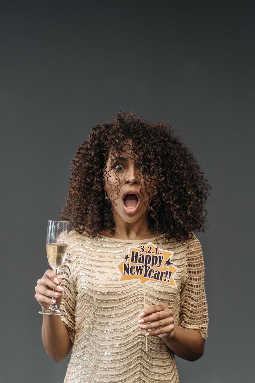 Astonished woman in a festive dress holding champagne and Happy New Year sign.