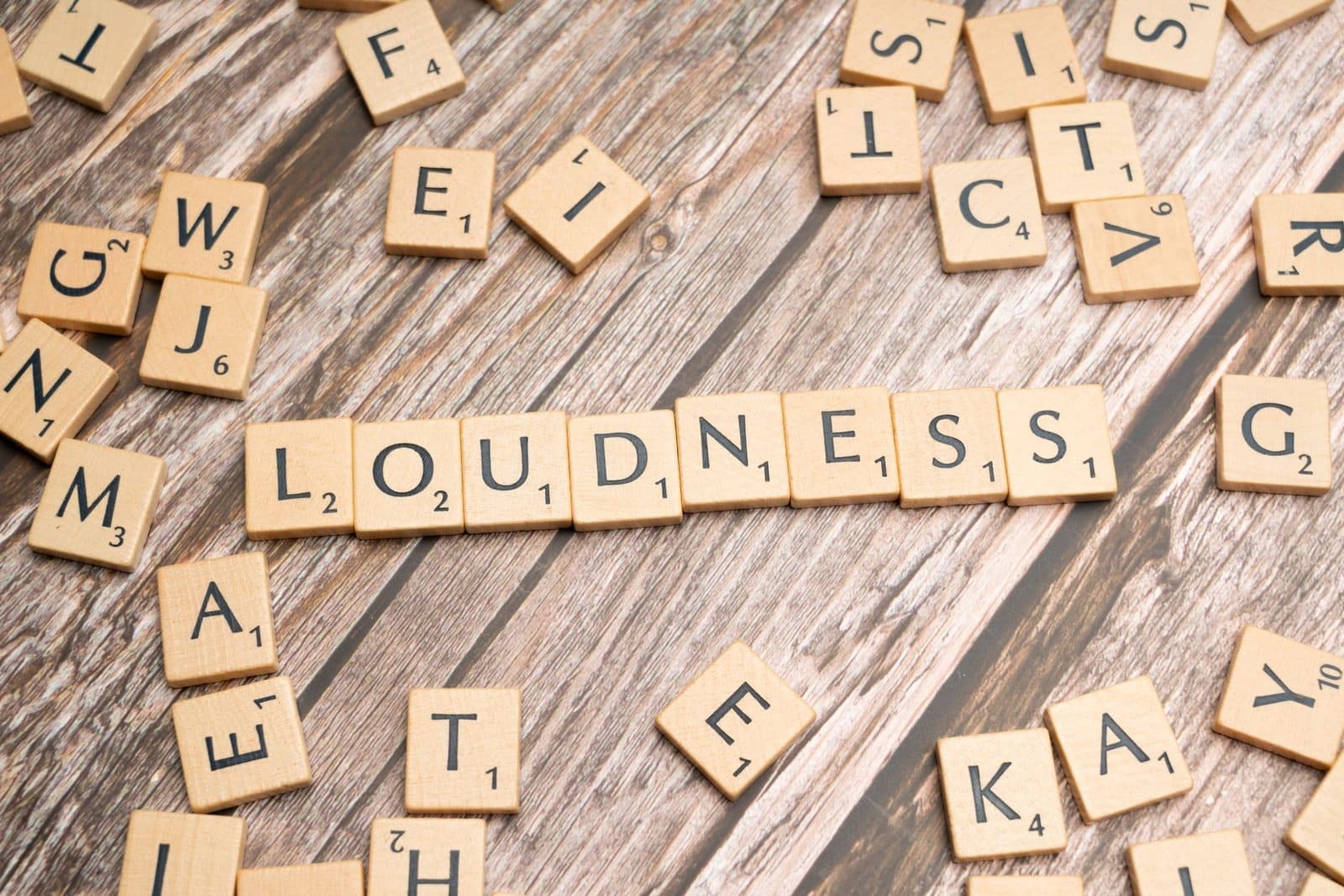 Scrabble tiles spelling 'Loudness' scattered over a wooden surface, emphasizing sound themes.