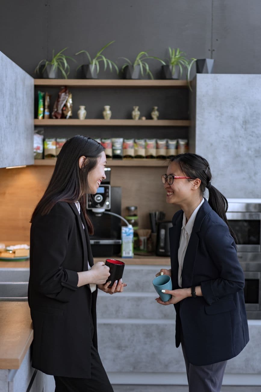 Two women in a modern office kitchen enjoying a coffee break, laughing and chatting.