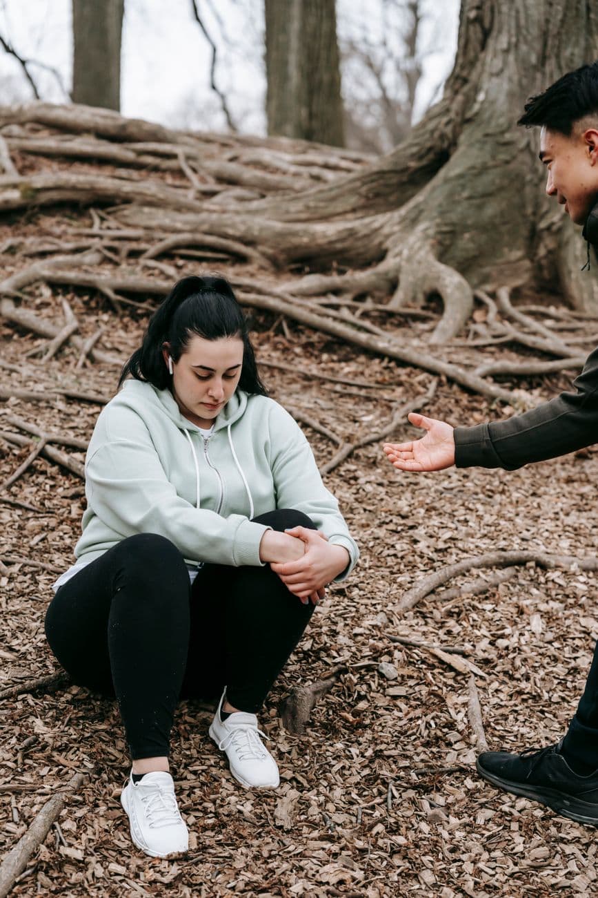 Crop positive young Asian man giving hand to serious offended girlfriend sitting on ground covered with fallen leaves during trekking in forest