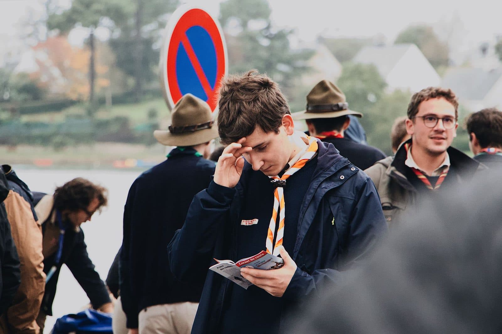 Young man in outdoor setting reading a brochure wearing winter clothes, with a no parking sign visible.