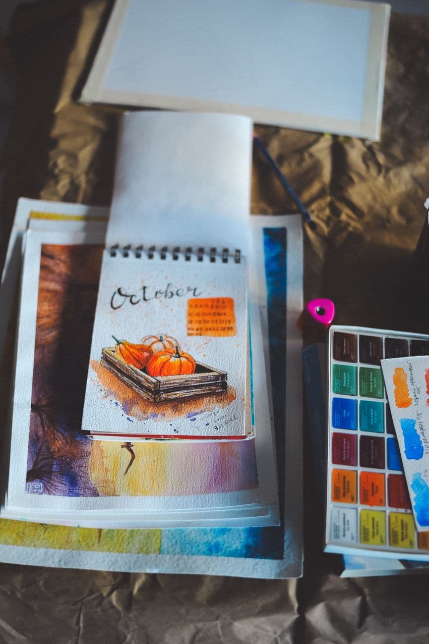 A sketchbook page with a watercolor pumpkin illustration and art supplies, embodying autumn creativity.