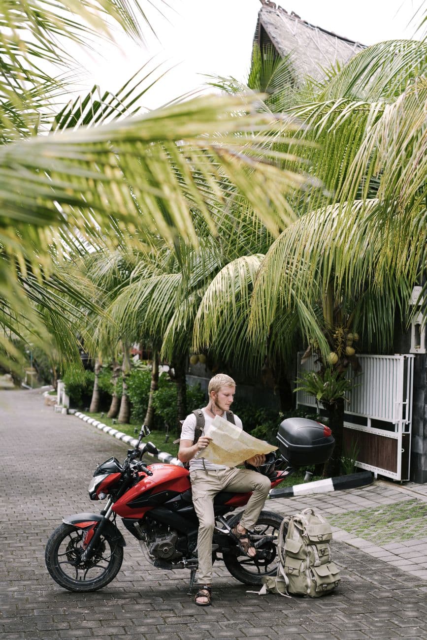 Full length male traveler studying map of city while leaning on motorcycle near duffle bag on street pavement among palms branches