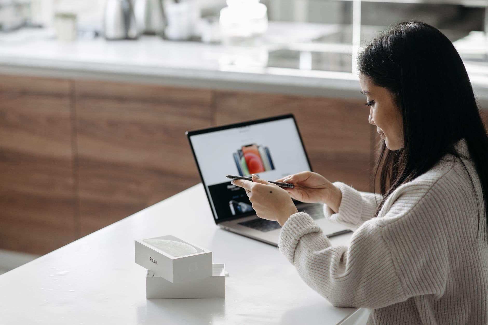 Young Asian woman setting up a new smartphone while working on a laptop in a modern indoor setting.