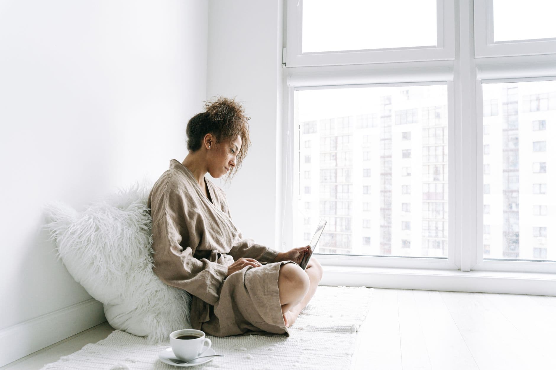 A woman in a robe sits by a window, using a laptop with a cup of coffee nearby.