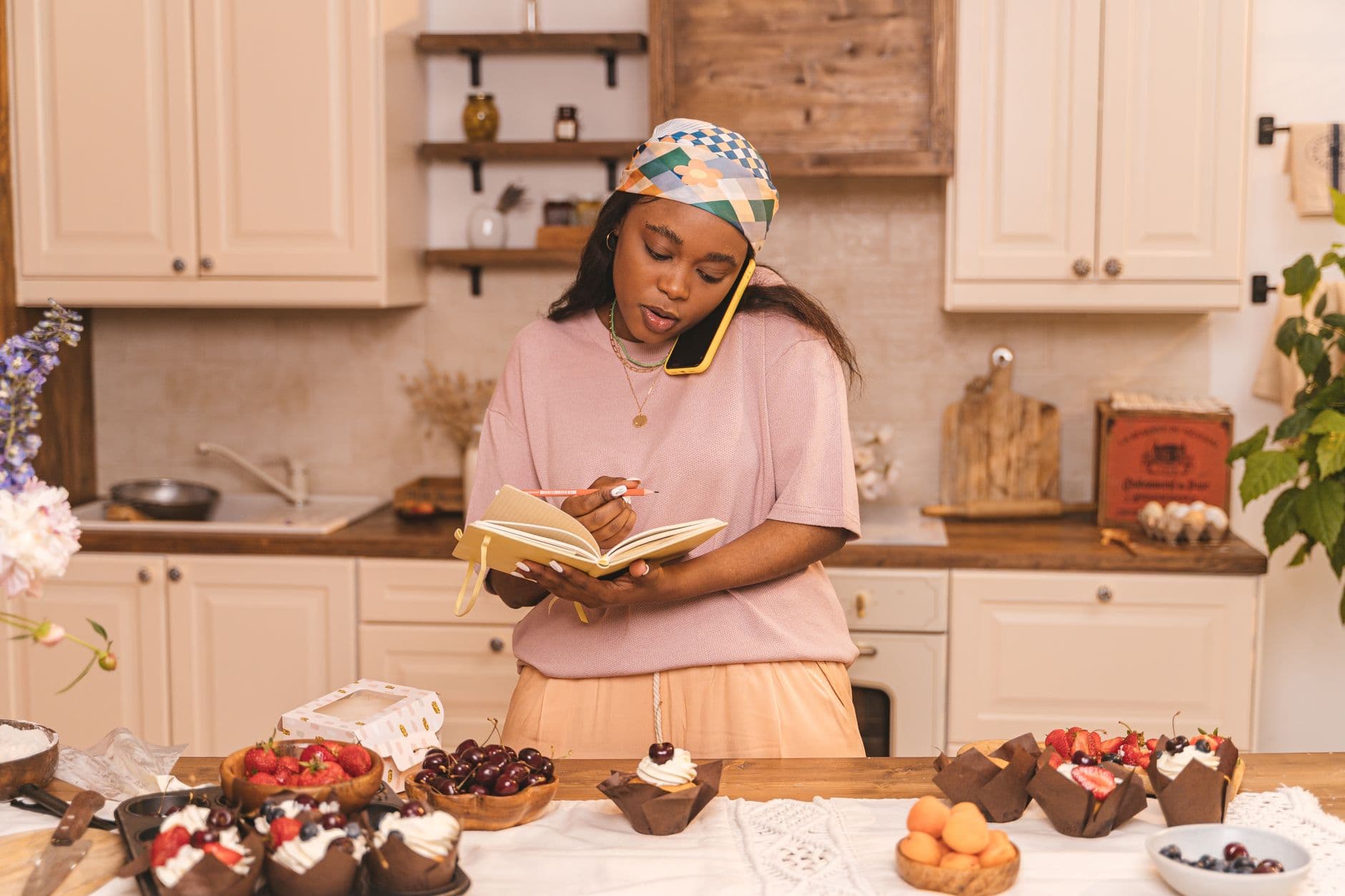 Black woman writing in notebook while on phone call surrounded by cupcakes in kitchen.