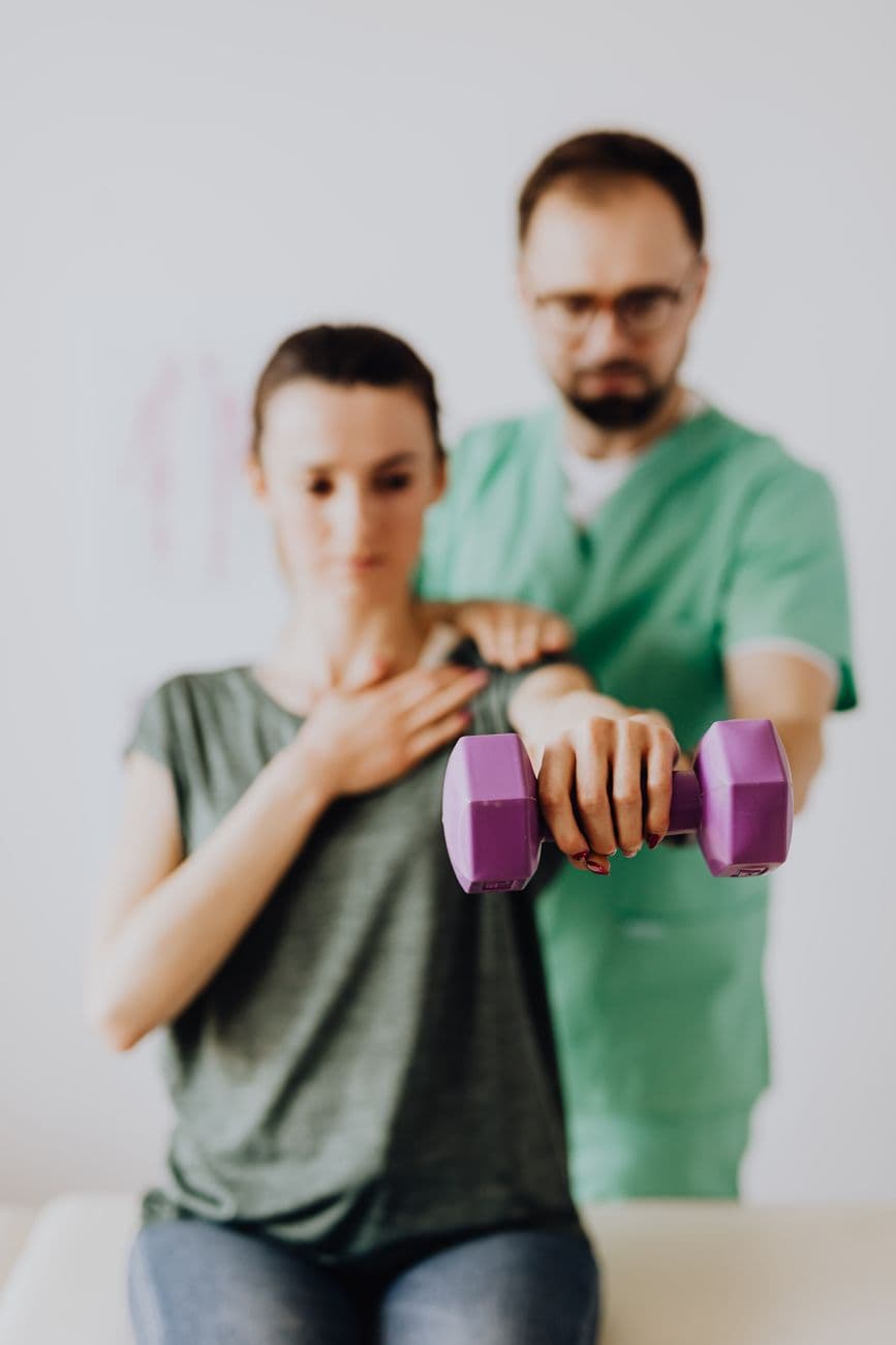 Bearded orthopedist in uniform and eyeglasses helping slim woman in casual clothes reaching arm with dumbbell while sitting on examination couch in clinic