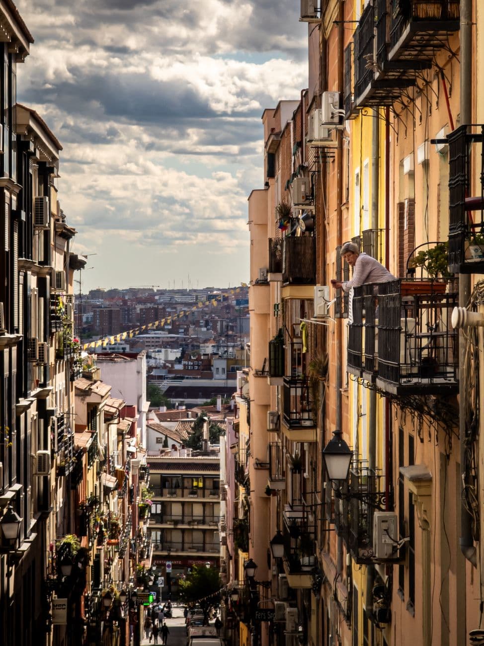A scenic street view of Madrid with residential buildings and a man on the balcony under dramatic skies.